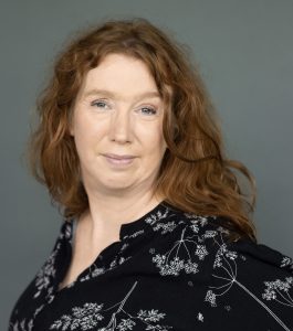 Portret van een vrouw met lange, krullende, rode haren, die glimlacht naar de camera. Ze draagt een zwarte blouse met een wit bloemenpatroon en staat tegen een effen achtergrond.