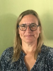 Portret van een vrouw met lang blond haar. Ze draagt een bril en kijkt met een glimlach. Ze staat voor een groene achtergrond.
